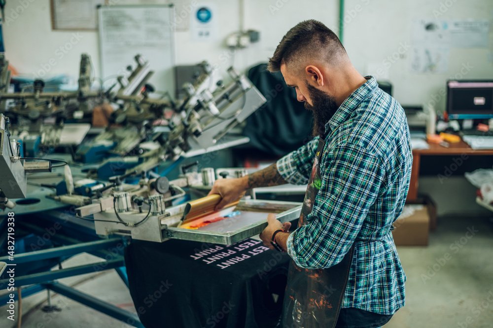 Male worker pressing ink on frame while using the printing machine in a workshop